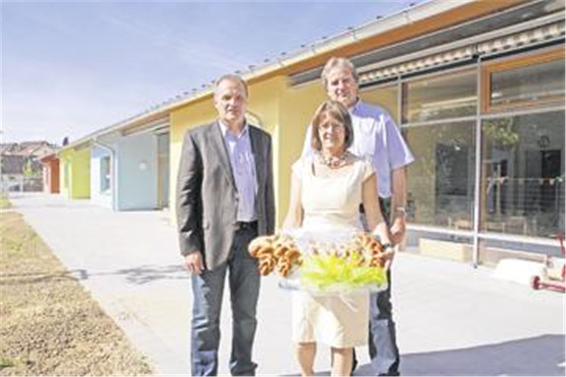 Architekt Martin Entenmann (li.) hat Kindergartenleiterin Marie-Luise Rohde zur Einweihung des im Hintergrund zu sehenden Anbaus den symbolischen Schlüssel in Gestalt von Hefegebäck mitgebracht. Darüber freut sich auch der Knittlinger Bürgermeister Heinz-Peter Hopp. Foto: Becker