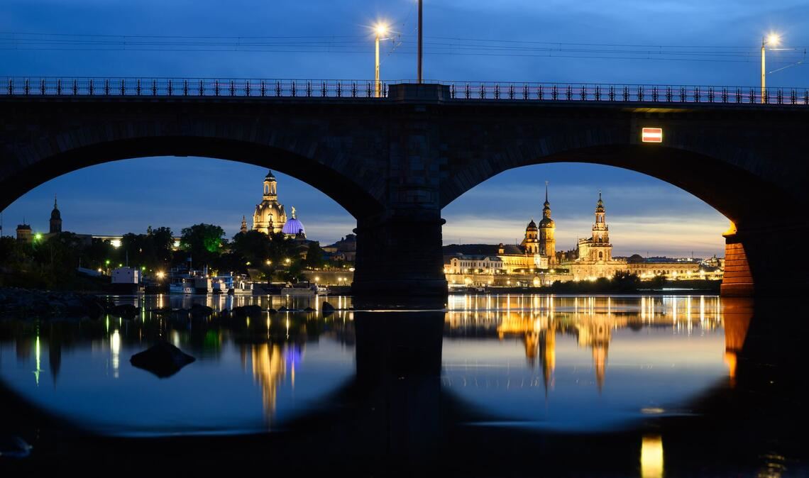 Blick am Abend in Dresden durch die Brückenbögen der Albertbrücke auf die historische Altstadtkulisse an der Elbe mit der Frauenkirche (l-r), der Kuppel der Kunstakedmie mit dem Engel „Fama“, dem Terrassenufer, dem Ständehaus, dem Hausmannsturm, der Hofkirche und der Semperoper.