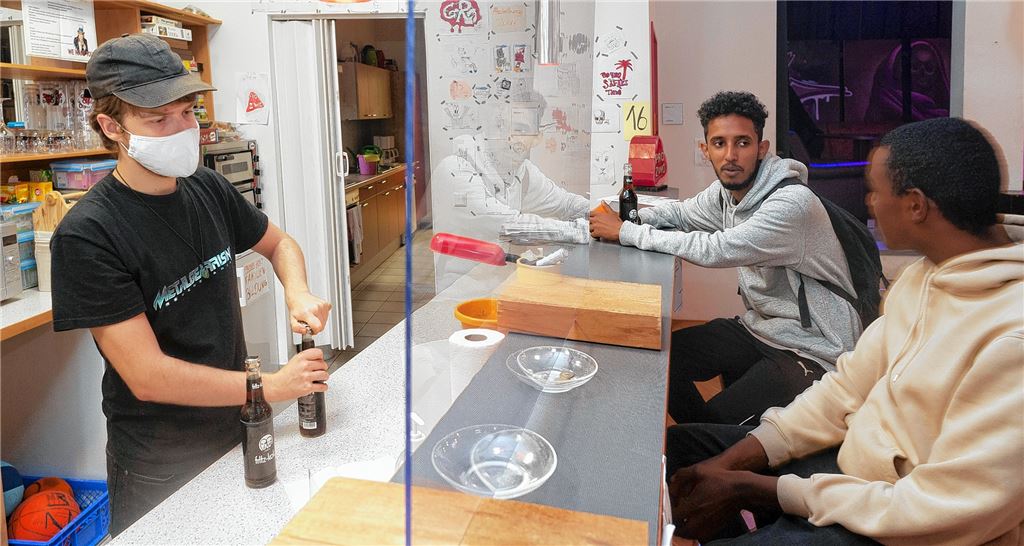 Bufdi Tobias bedient mit Mundschutz und hinter einer Plexiglasscheibe, Travis im grauen Oberteil und Tupac sind trotz Corona-Einschränkungen am Samstagabend im Jugendhaus.Fotomoment