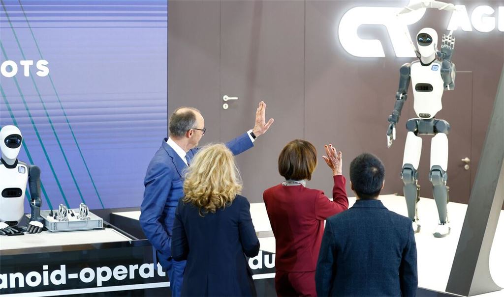 Bundeskanzler Friedrich Merz winkt einem Roboter zu – die Zukunft winkt zurück.