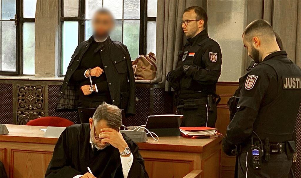 Der Angeklagte (hinten links) steht in Handschellen im Verhandlungssaal im Landgericht neben Justizvollzugsbeamten.