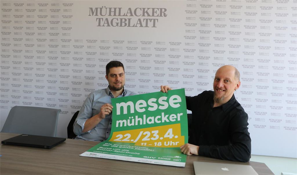 Messeorganisatoren von der Messe Mühlacker Alexander Kachur und Björn Grau zeigen das Werbeplakat zur Messe. Auf diesem Steht drauf, dass die Messe Mühlacker am 22. und 23. April von 11 bis 18 Uhr stattfindet. Im Hintergrund sieht man das Logo der Tageszeitung Mühlacker Tagblatt.