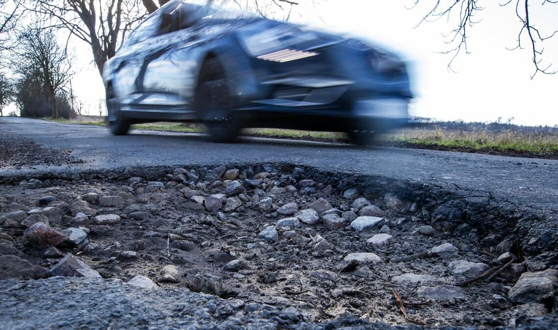 Ein Auto fährt an einem Schlagloch in einer Allee vorbei. Das Frostwetter der letzten Wochen sorgt für kaputte Straßen mit tiefen Schlaglöchern.
