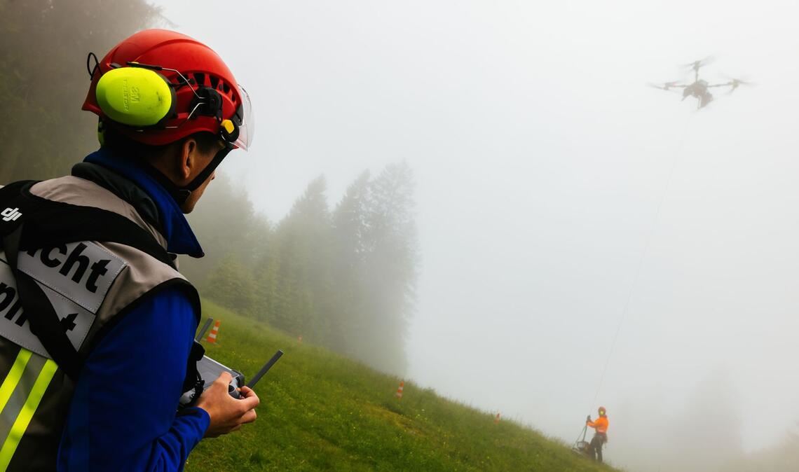 Ein Drohnenpilot der Bergwacht sieht zu, wie ein Flughelfer der Bergwacht eine Übungslast von der Seilwinde einer Lastendrohne abhängt.