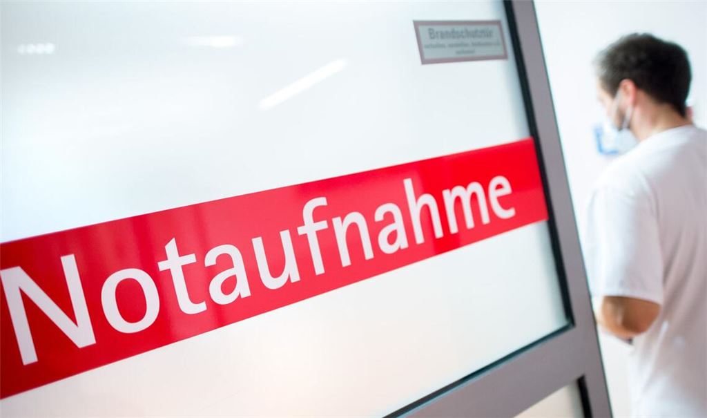 Ein Krankenpfleger steht in einem Krankenhaus neben einer Glastür mit der Aufschrift „Notaufnahme“.