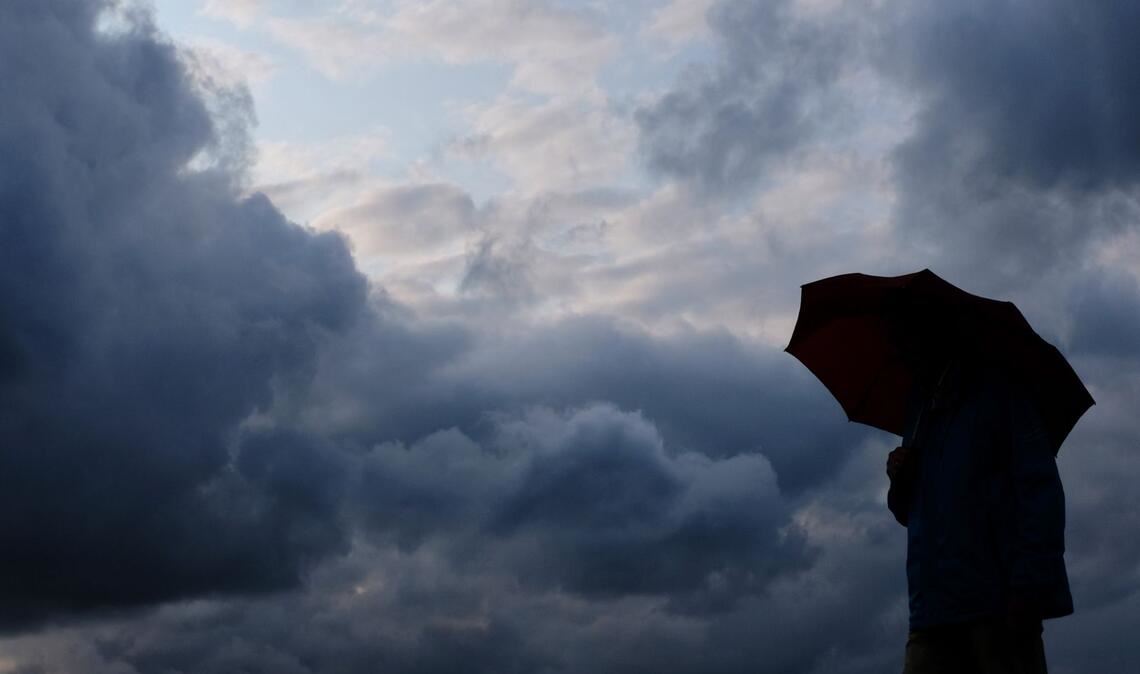 Ein Mann geht mit einem Regenschirm vor aufziehenden dunklen Wolken spazieren. (Symbolbild)
