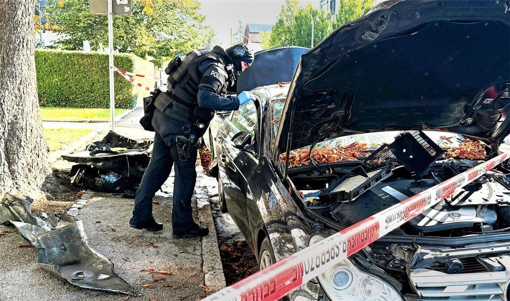 Ein Polizist mit Schutzweste steht an einem PKW, der gebrant hatte. Ein Brand in einem Münchner Einfamilienhaus hat einen Großeinsatz von Polizei und Feuerwehr ausgelöst.