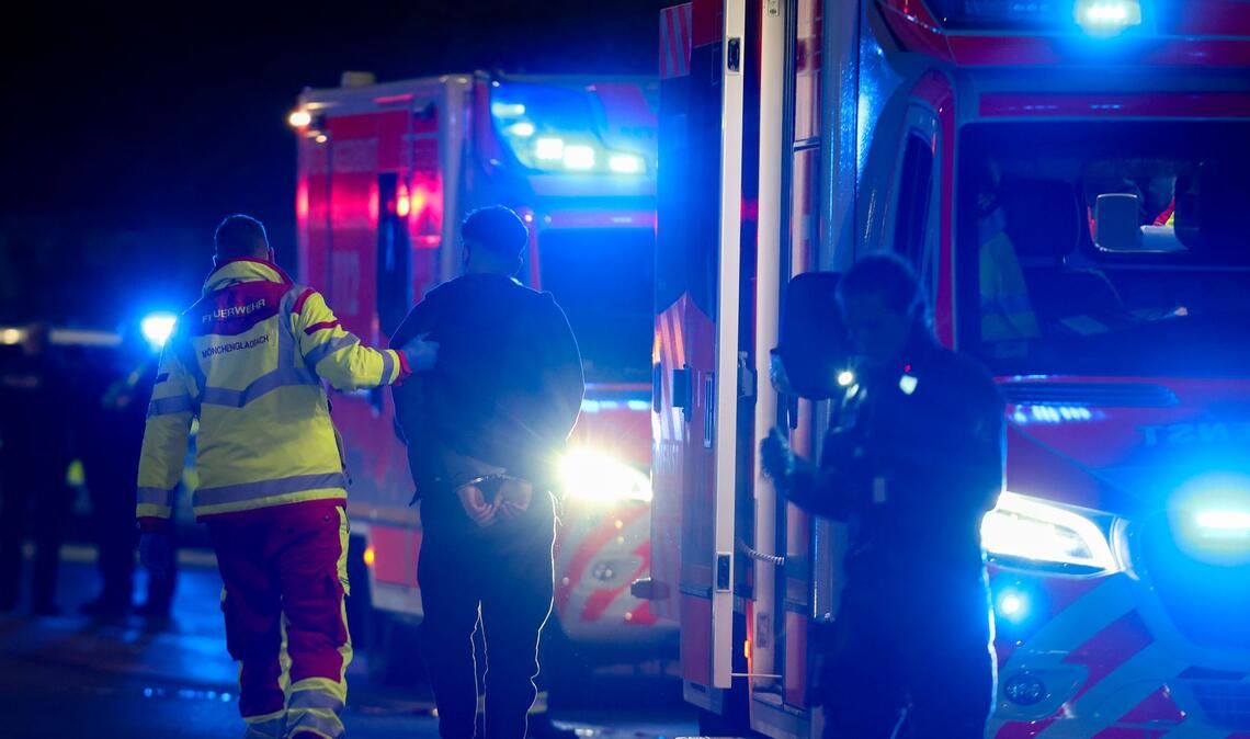 Ein Tatverdächtiger in Handschellen wird nach Schüssen in Mönchengladbach von einem Sanitäter zu einem Rettungswagen gebracht.