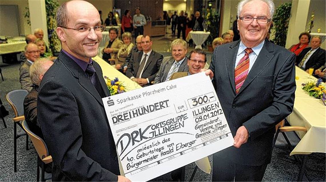 Ein strahlender Jubilar Harald Eiberger (li.) nimmt von Winfried Scheuermann einen Spendenscheck für das DRK Illingen entgegen. Im Hintergrund: eine Schar fröhlicher Gäste.