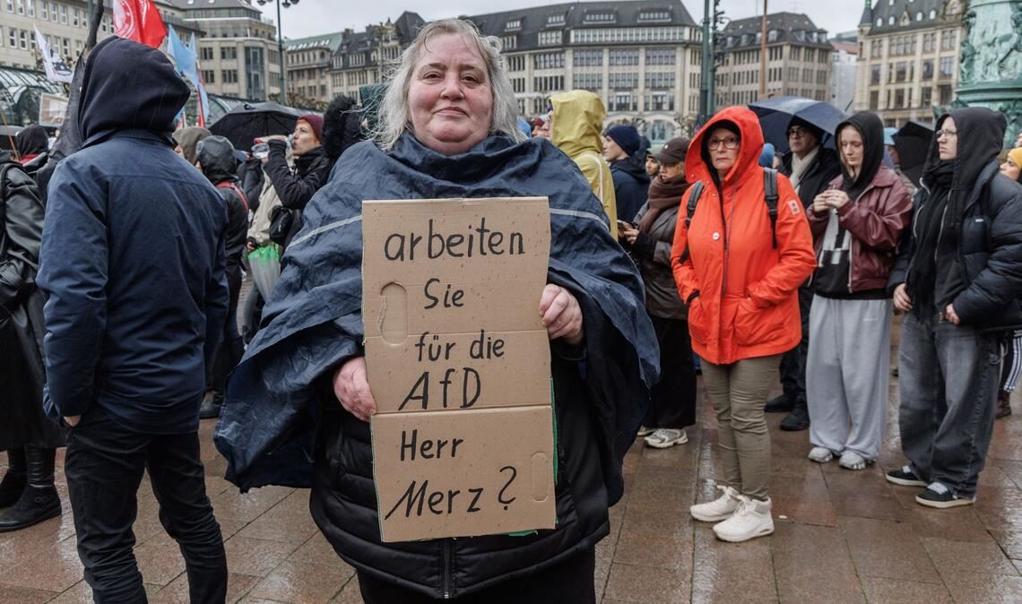 Eine Demonstrantin in Hamburg stellt auf einem Pappschild eine Frage an Bundeskanzler Friedrich Merz (CDU).
