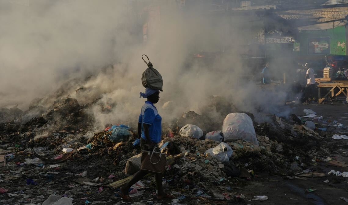 Eine Frau, die eine Tasche auf dem Kopf trägt, geht im Stadtteil Petion-Ville von Port-au-Prince an schwelendem Müll vorbei.