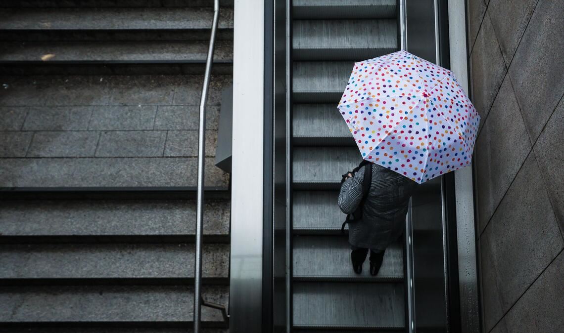 Eine Frau fährt mit ihrem Regenschirm eine Rolltreppe hinauf. Das Wetter soll in der Region Stuttgart in den nächsten Tagen wechselhaft bleiben.