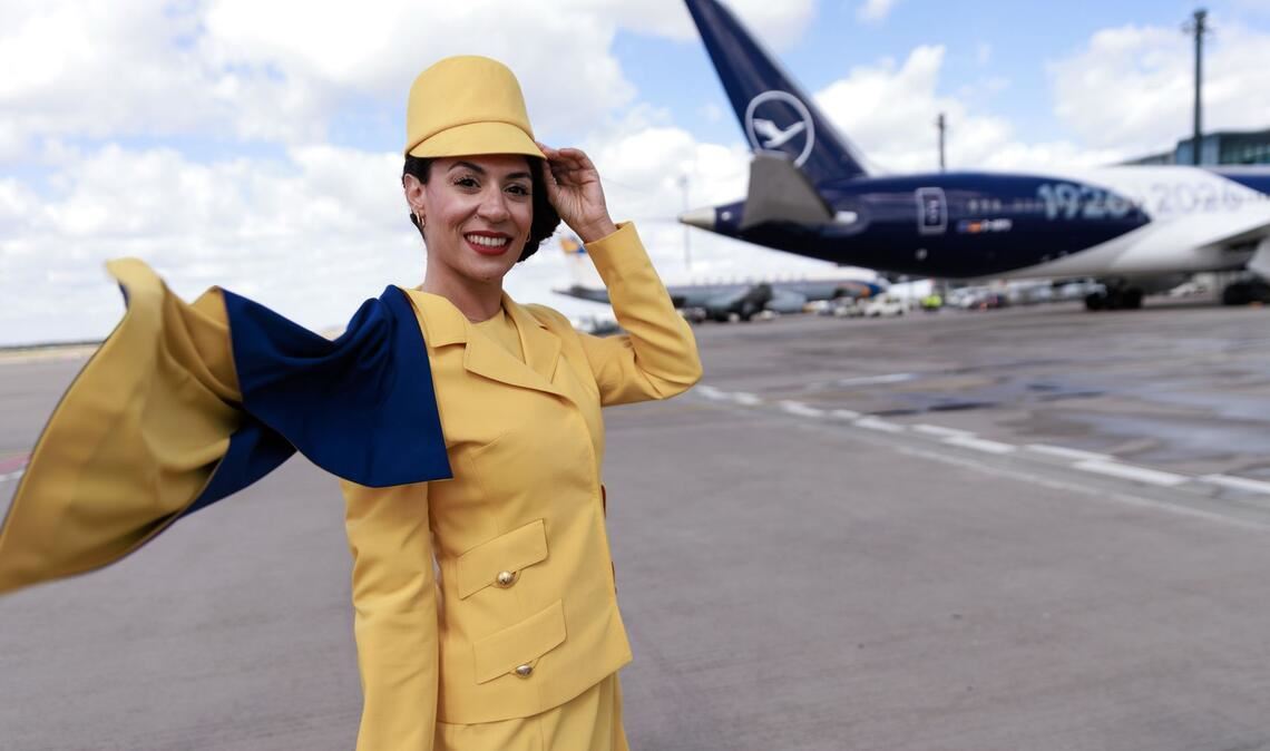 Eine Hostess trägt eine frühere Lufthansa-Uniform vor einem Passagierflugzeug der Lufthansa auf dem Flughafen Berlin-Brandenburg (BER) bei der Feier von Lufthansa zum 100. Jahrestag des Erstflugs.