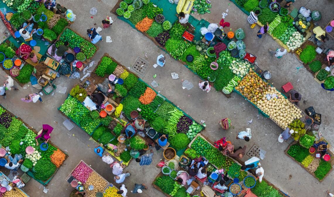 Eine Luftaufnahme zeigt das lebhafte Treiben auf einem Gemüsemarkt in Ahmedabad in Indien, wo Verkäufer frische Produkte anordnen und Kunden sich durch die engen Marktgassen bewegen.