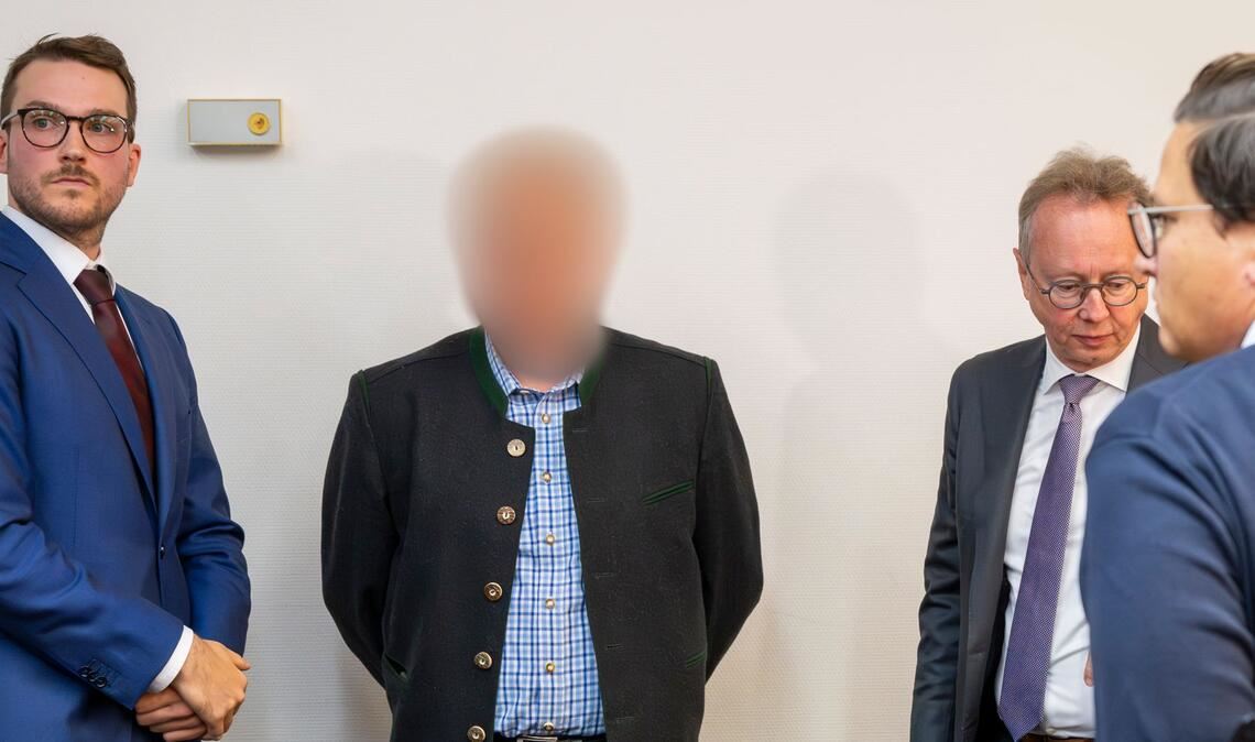 Einer der beiden beschuldigten Bahnmitarbeiter (M) steht vor Prozessbeginn zwischen Anwälten im Gerichtssaal des Landgerichts München.