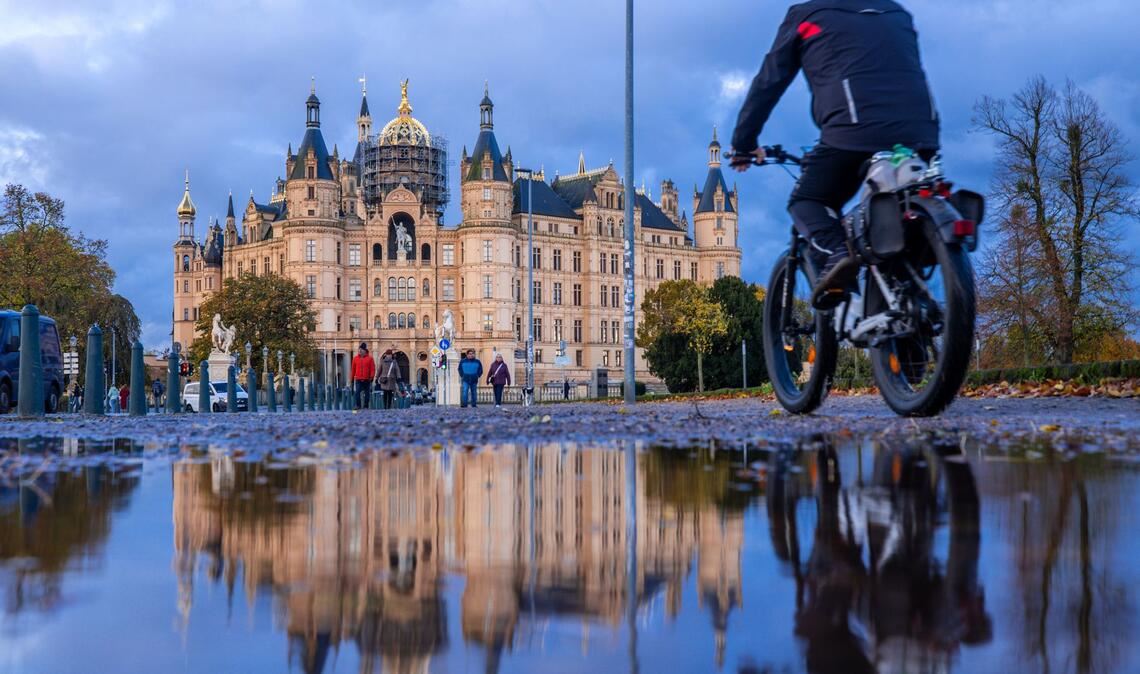 Herbstwetter in Norddeutschland: Ein Radfahrer fährt an einer Pfütze vorbei, in der sich das Schweriner Schloss spiegelt.