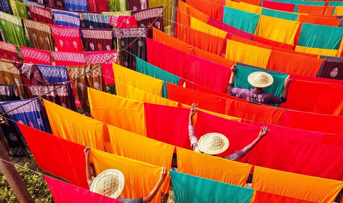 In einer Färberei in Bangladesch hängen unzählige gefärbte Stoffbahnen zum Trocknen bei Temperaturen von über 42 Grad Celsius. Die Arbeiter und Arbeiterinnen haben dabei allerhand zu tun: Sie müssen die Stoffe ständig drehen.
