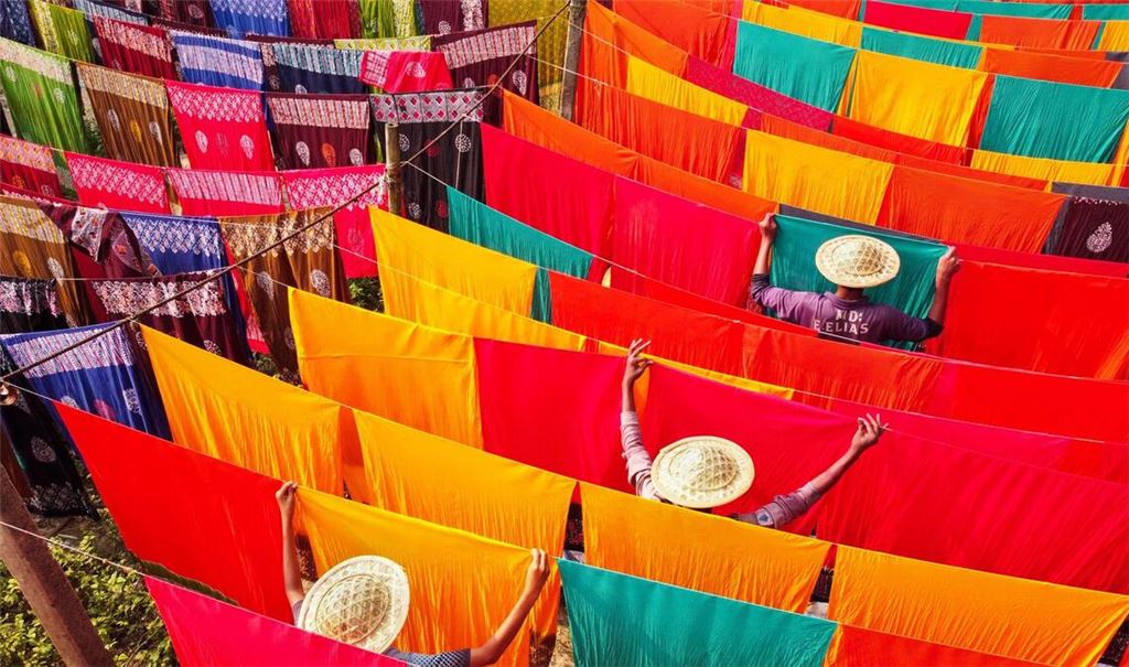 In einer Färberei in Bangladesch hängen unzählige gefärbte Stoffbahnen zum Trocknen bei Temperaturen von über 42 Grad Celsius. Die Arbeiter und Arbeiterinnen haben dabei allerhand zu tun: Sie müssen die Stoffe ständig drehen.