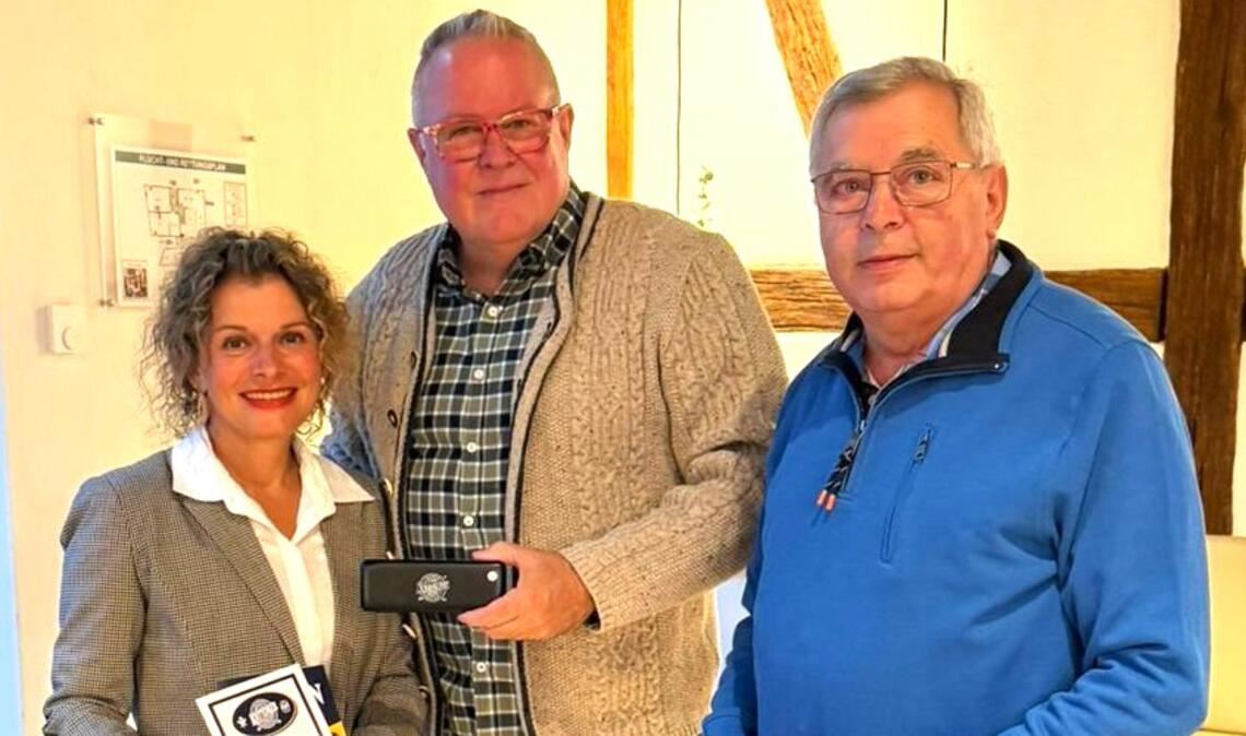 John Hall überreicht seine „Knittlinger“ als Gastgeschenk an die stellvertretende Bürgermeisterin Patricia Lopez. Rechts Manfred Dannecker, Dirigent des Mundharmonika-Orchesters Knittlingen. Fotos: Haller