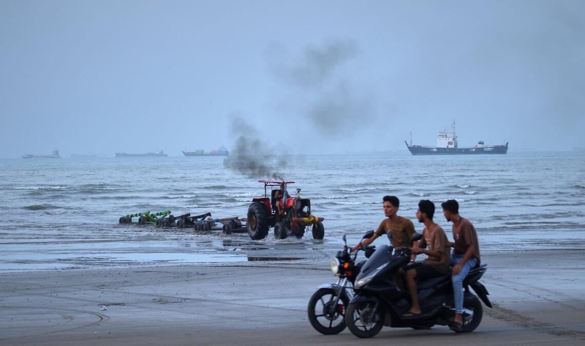Junge Männer fahren mit Motorrädern am Ufer entlang, während ein Traktor Geräte aus dem Wasser zieht und Handelsschiffe in der Straße von Hormuz vor Anker liegen. (zu dpa: «Rund 400 Schiffe stecken noch im Persischen Golf fest»)