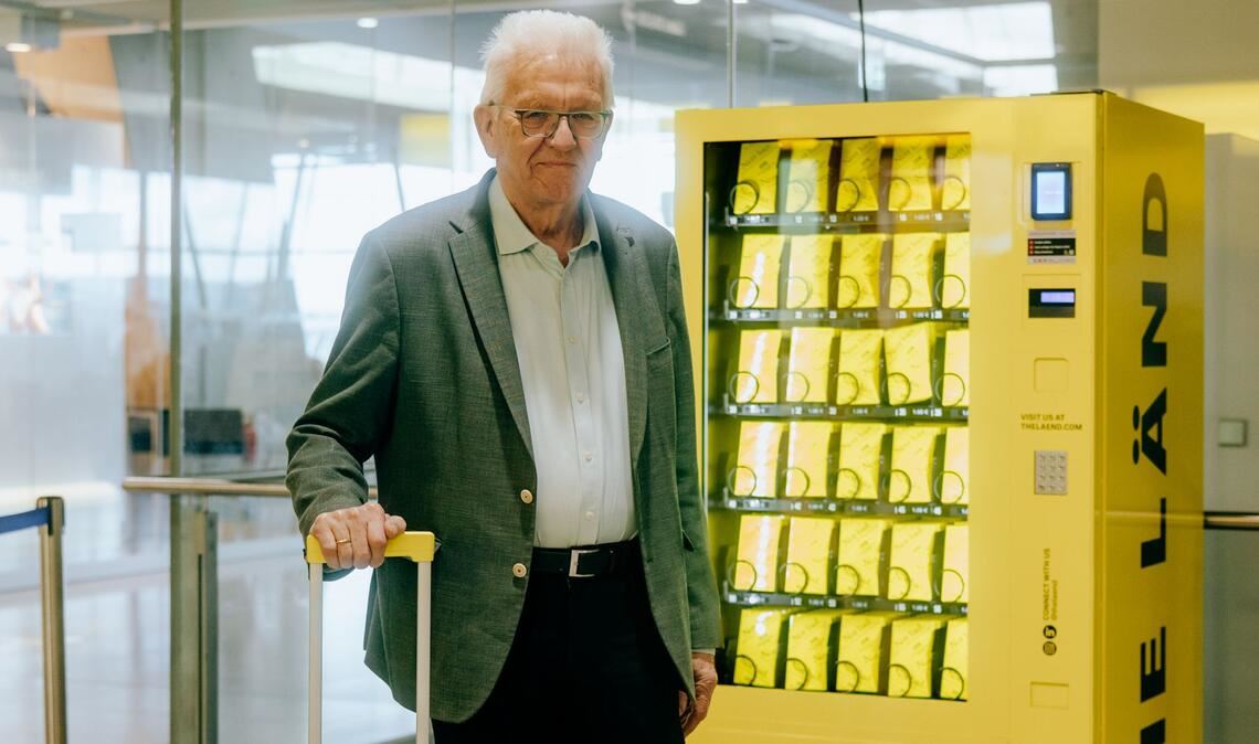 Ministerpräsident Winfried  Kretschmann vor dem „Nett-hier“-Sticker-Automat am Flughafen.