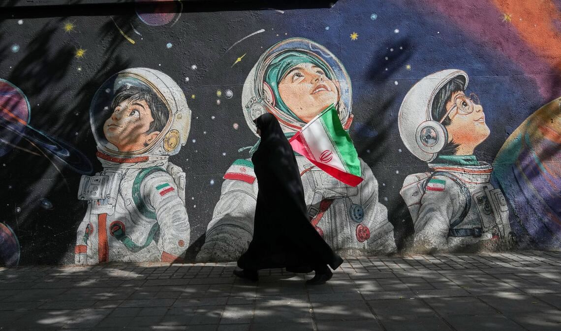 Mit einer iranischen Flagge in der Hand geht eine Frau an einer Wand vorbei, auf der Kinder in Raumanzügen zu sehen sind.