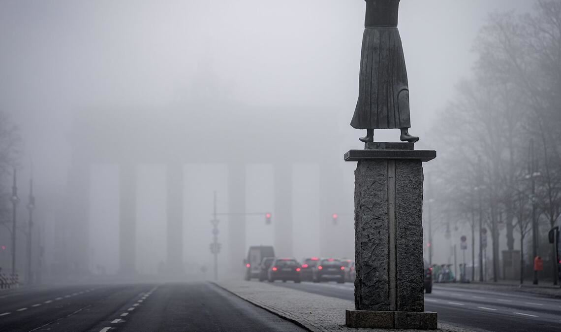 Schemenhaft ist das Brandenburger Tor im Nebel im Hintergrund der Bronzefigur "Der Rufer" von Gerhard Marcks am frühen Morgen zu sehen.
