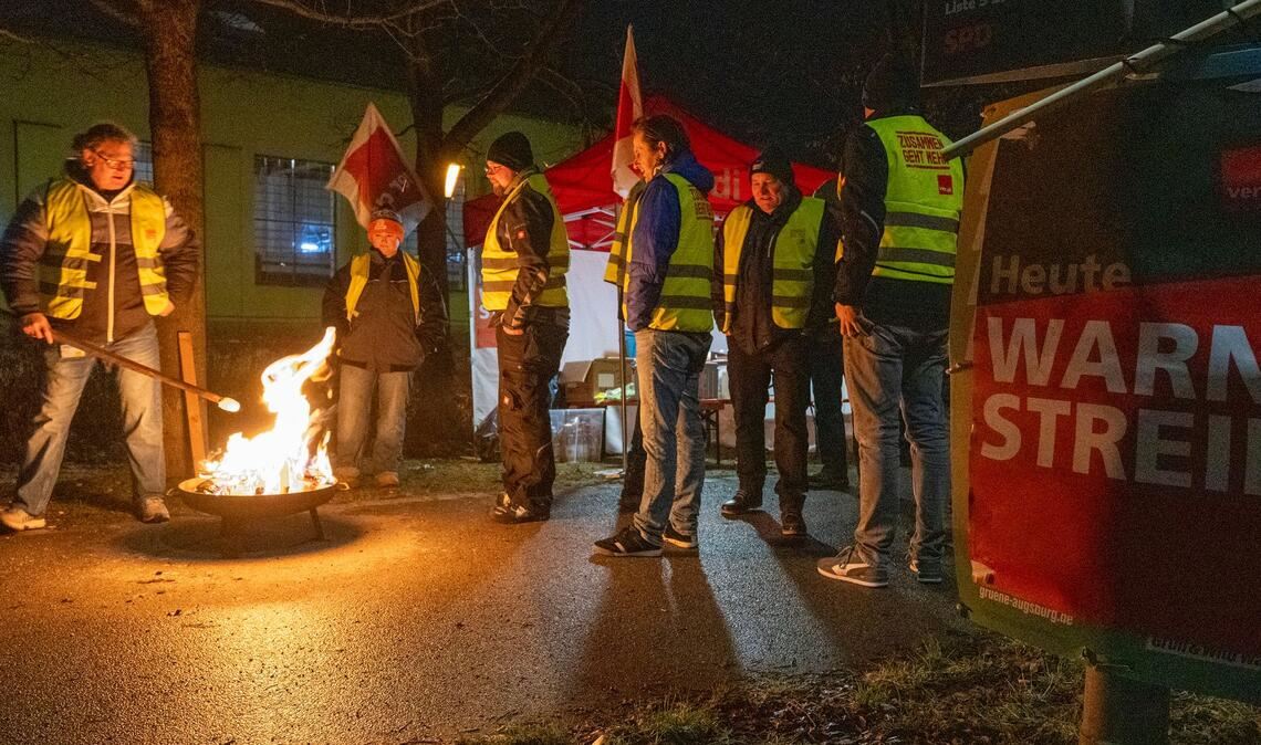 Streikende in Warnwesten wärmen sich in Augsburg an einem Feuer während eines Warnstreiks im ÖPNV.