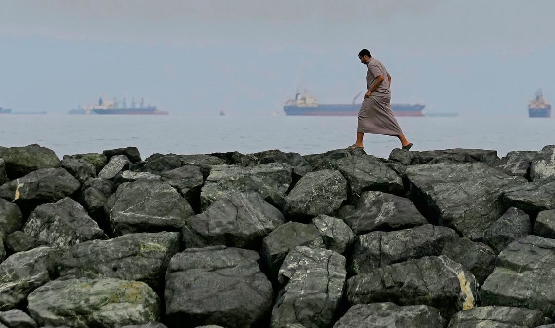 Vereinigte Arabische Emirate, Chaur Fakkan: Ein Mann geht auf den Felsen am Ufer spazieren, während Öltanker und Frachtschiffe in der Straße von Hormus aufgereiht sind.