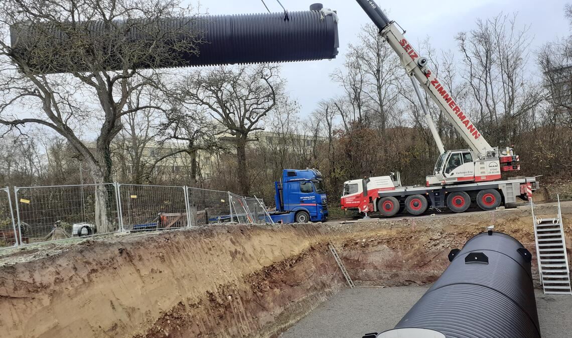 Während eine der beiden Löschwasserzisternen bereits an Ort und Stelle liegt, wird die andere mit einem mobilen Kran in die Baugrube gehievt. Foto: Gezener