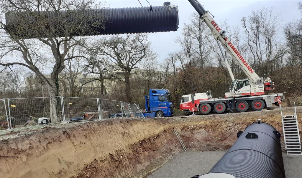 Während eine der beiden Löschwasserzisternen bereits an Ort und Stelle liegt, wird die andere mit einem mobilen Kran in die Baugrube gehievt. Foto: Gezener