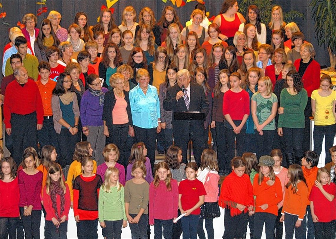 Zusammen mit rund 100 Sängerinnen und Sängern des DIG und des Theodor-Heuss-Gymnasiums auf der herbstlich geschmückten Bühne begrüßt DIG-Vorsitzender Hermann Fasching (Mitte) mit launigen Worten die Zuhörer im ausverkauften Saal des Uhlandbaus. 